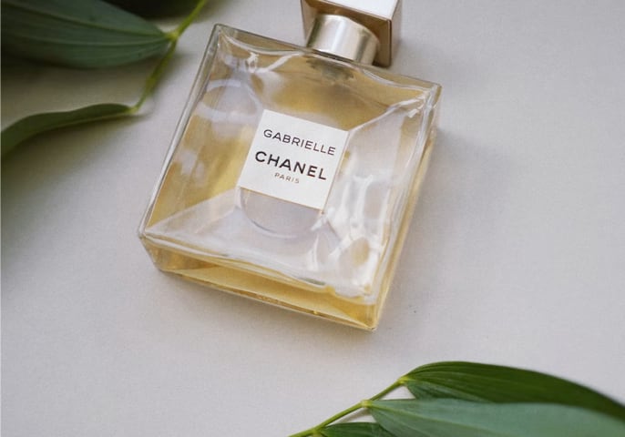 Bottle of Gabrielle Essence Eau De Parfum laying on a white background next to leaves